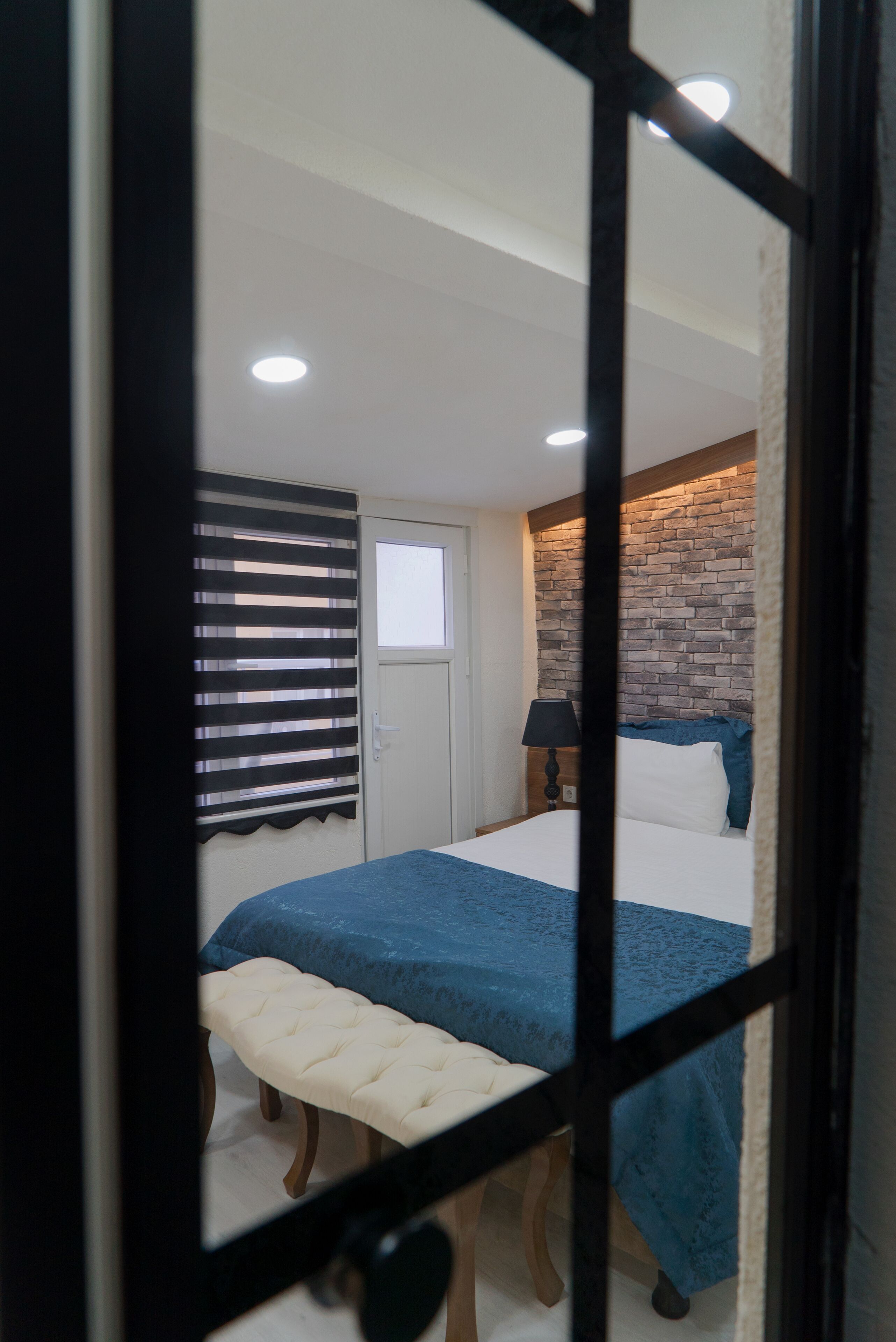A bedroom with a bed, a bench, a window with blinds, a door, and a brick-patterned wall.