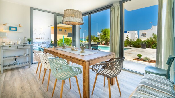 A modern dining area with a wooden table and chairs, large windows with a view of the sea and buildings, and a hanging light fixture. - Kasalmar, Beach House 5StarsHome Mallorca, private swimming pool,air,wifi,beach. (Son Serra de Marina)