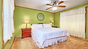 Une chambre à coucher comprenant un lit, deux tables de chevet, un ventilateur de plafond, un élément de décoration murale et une fenêtre avec des rideaux.