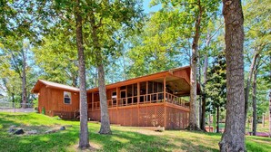 Une cabane en bois dotée d’un balcon, entourée d’arbres et bordée d’une zone herbeuse.