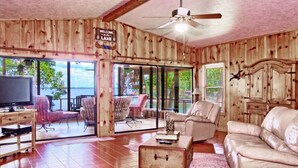 Une cabane avec un intérieur en bois, un ventilateur de plafond, une télévision, un canapé, une table basse et une vue sur le lac.