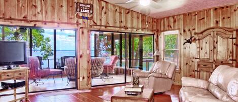 A cabin with a wooden interior, a ceiling fan, a television, a sofa, a coffee table, and a view of the lake.