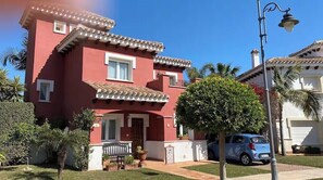 Une maison à deux étages avec une façade rouge, un balcon et une voiture garée dans l’allée.