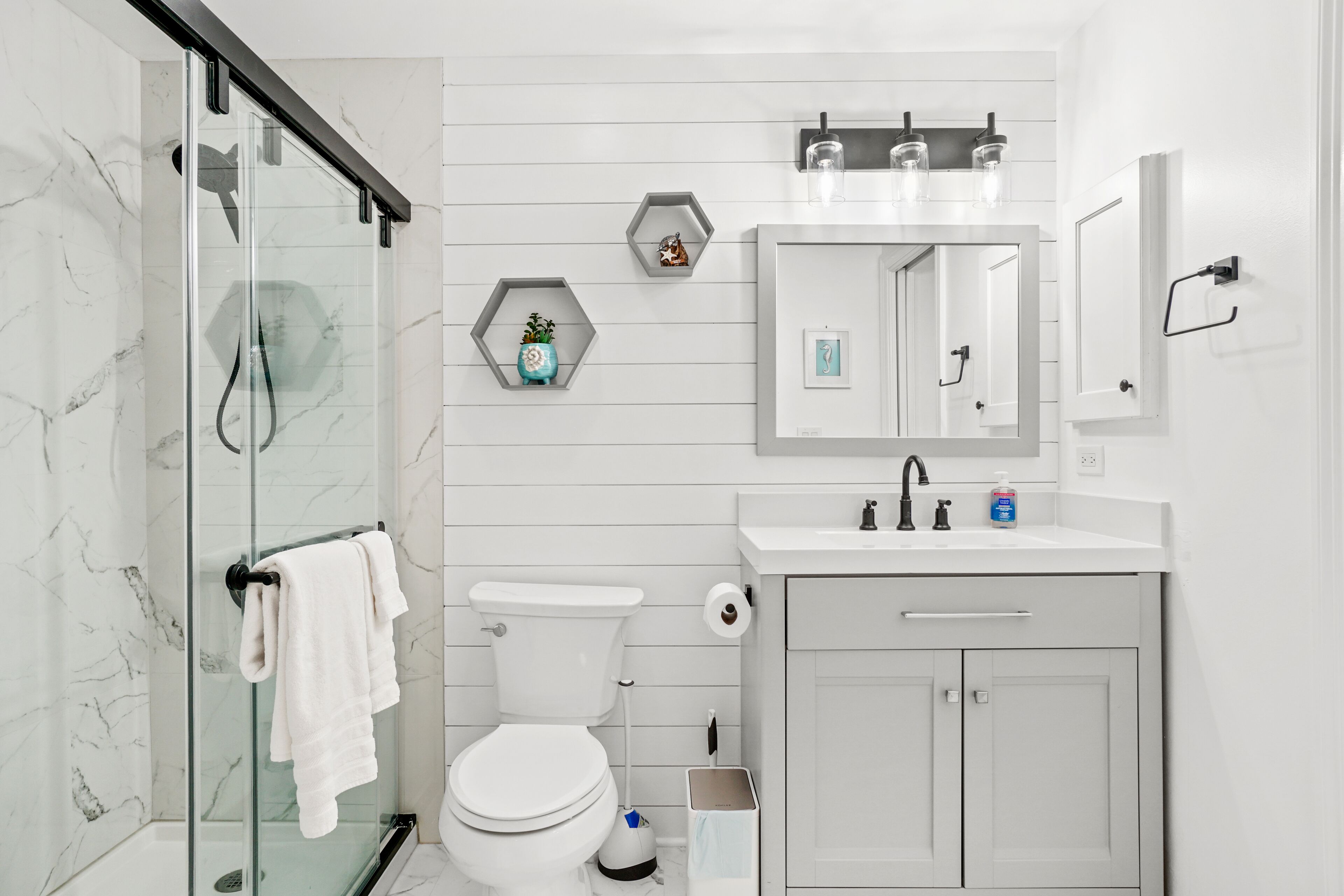Une salle de bain moderne avec une douche à parois de verre, des toilettes, un lavabo avec un miroir au-dessus et une petite étagère ornée d’objets décoratifs.