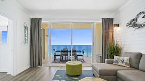 Un salón con sofá, reposapiés y un control remoto sobre una mesa. Un ventanal con cortinas da a un balcón con vistas al océano.