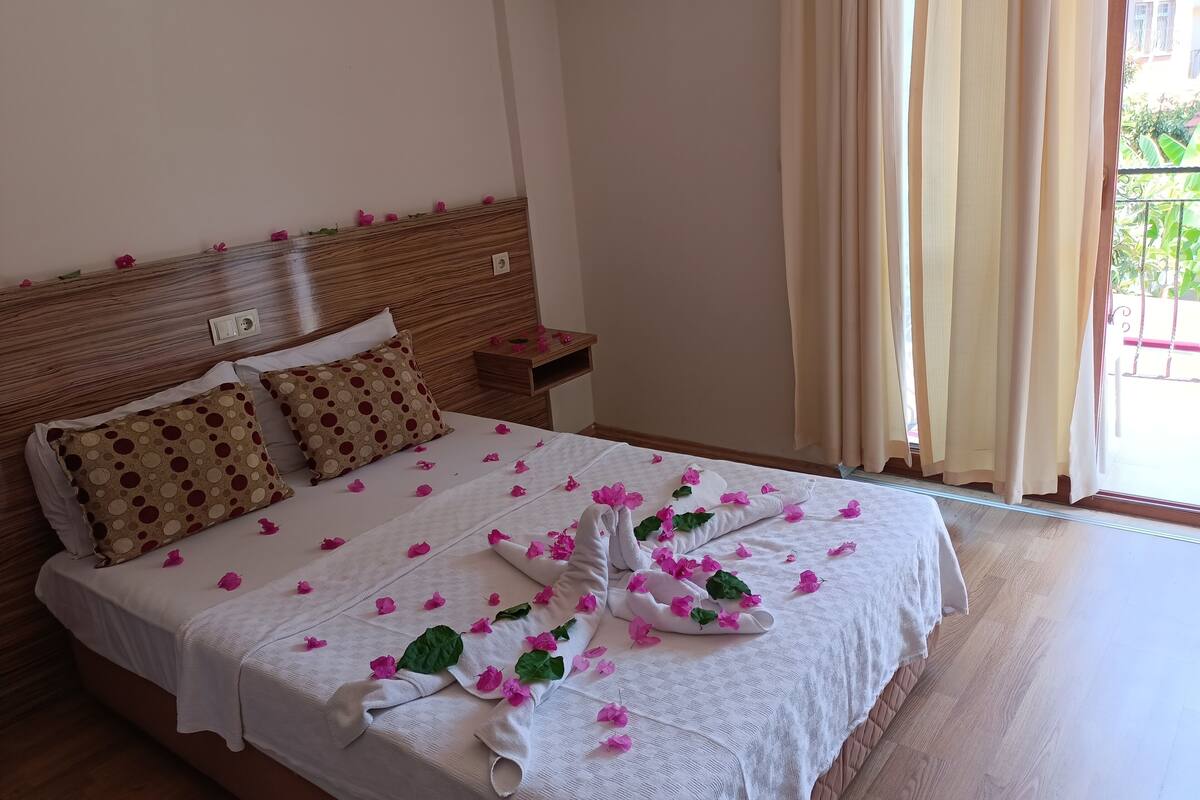 Una cama bien hecha con sábanas blancas y pétalos de flores rosadas, un cabecero de madera y una mesita de noche pequeña.