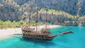 A large, multi-decked ship with multiple masts and a dragon figurehead, docked at a beach with clear turquoise waters and a rocky backdrop. - Hotel Barlas Fethiye (Fethiye)