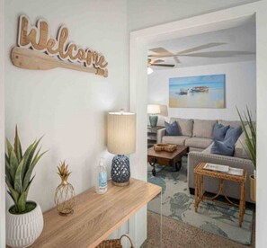 Un salon avec une table basse en bois, un canapé, une lampe et un panneau mural sur lequel on peut lire « Bienvenue ».