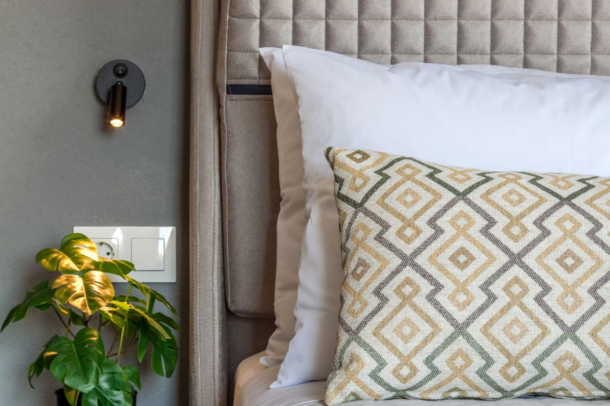 Un lit bien fait, agrémenté d’un coussin à motifs, d’une lampe de chevet et d’une plante en pot posée sur une table de chevet.