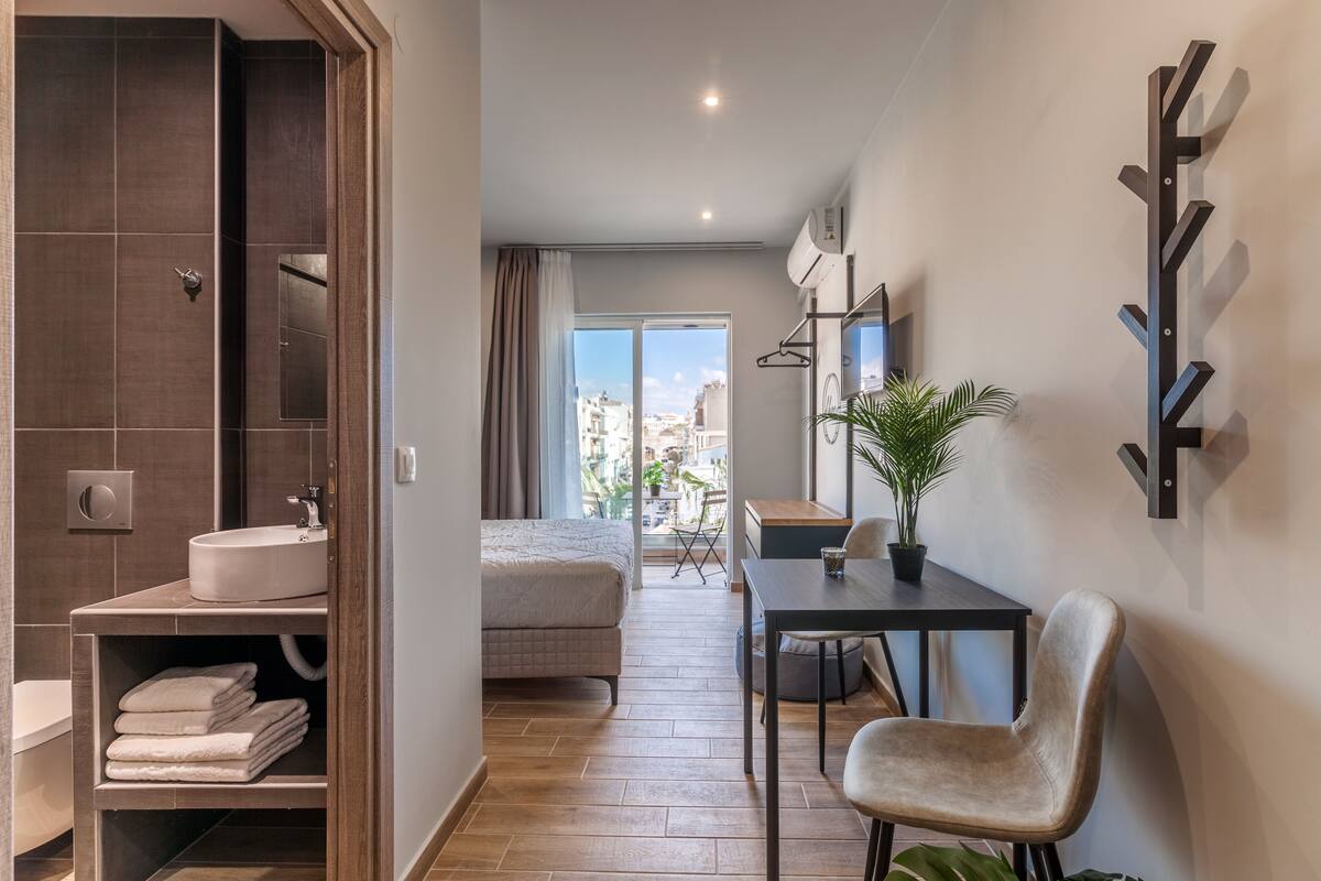 Une chambre d’hôtel moderne avec un grand lit, une petite table avec une chaise, une plante en pot et une vue sur la ville par la fenêtre.