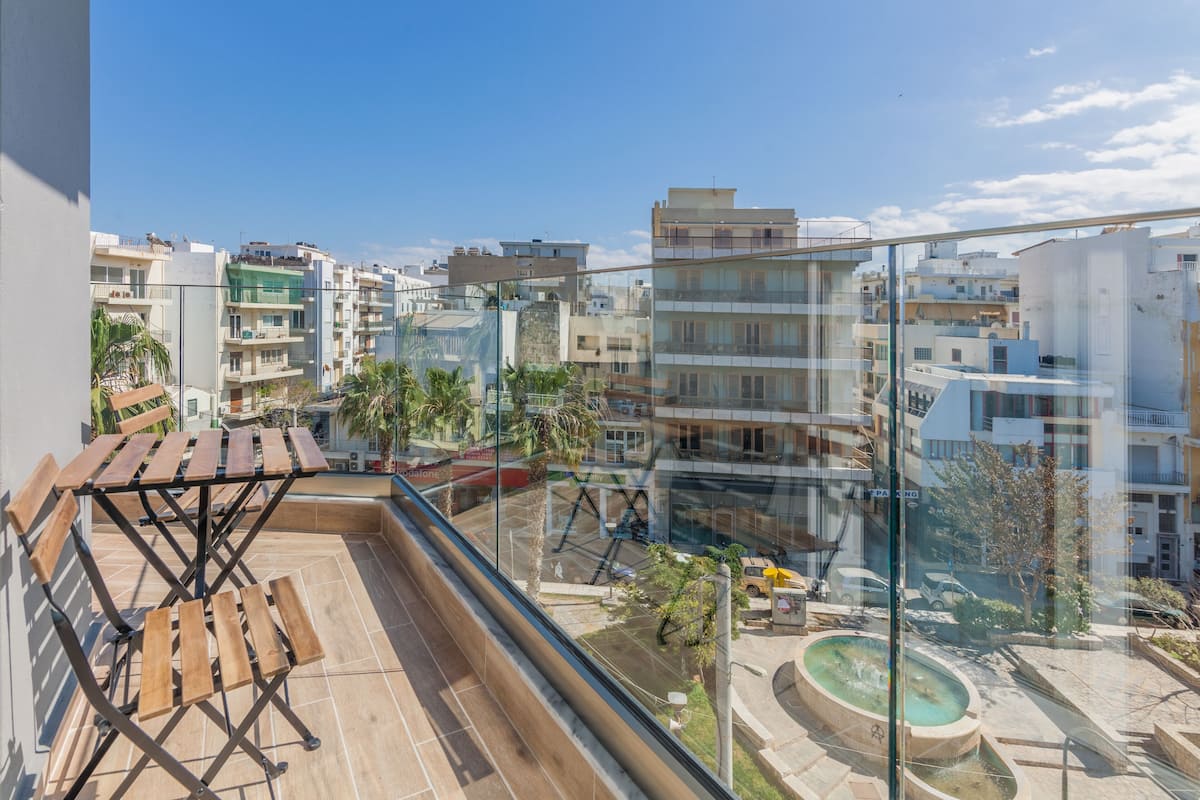 Un balcon avec une table et des chaises en bois, donnant sur un paysage urbain composé d’immeubles modernes et d’une fontaine.