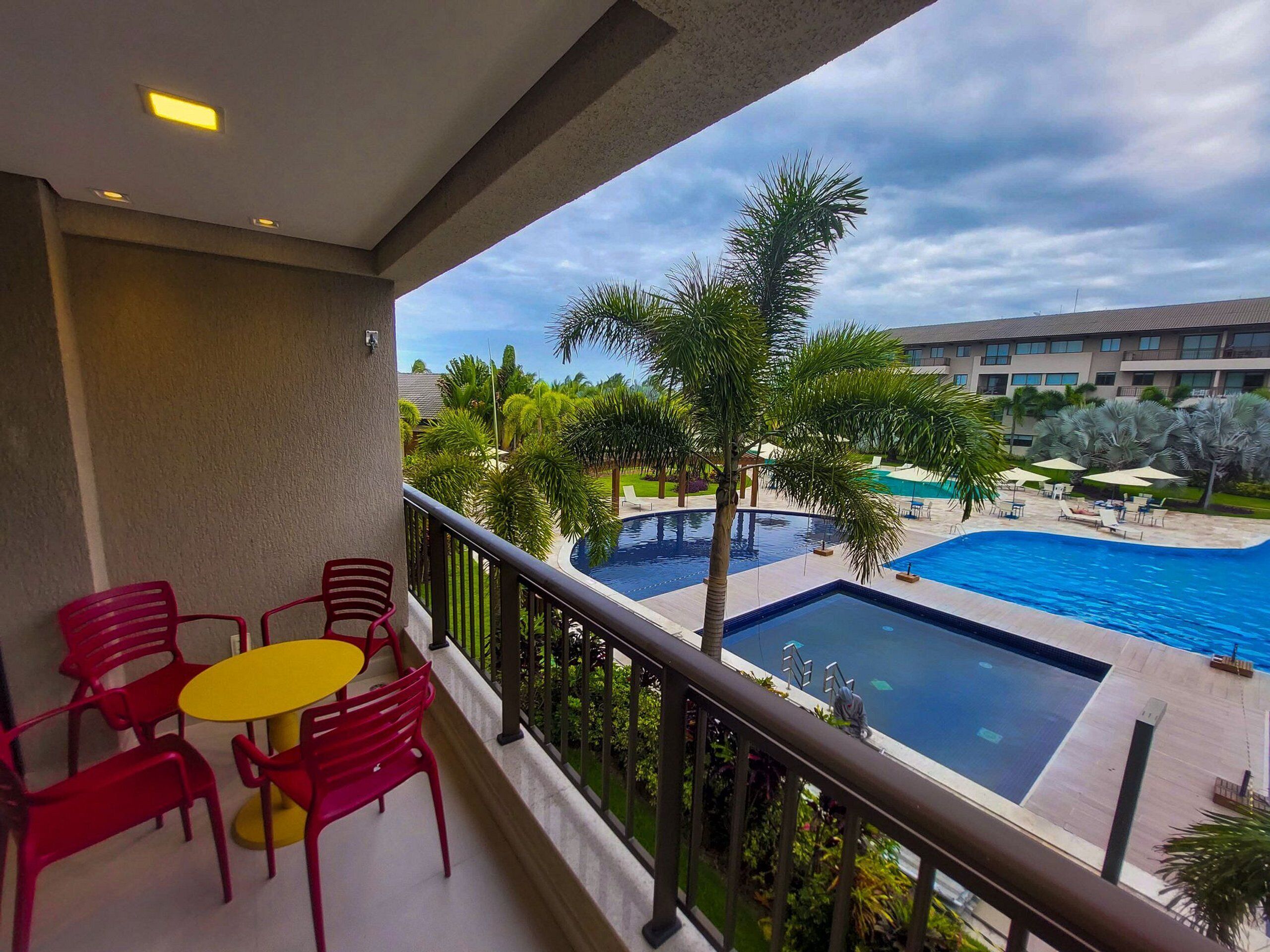 A balcony with red chairs and a yellow table, overlooking a swimming pool and a building with multiple floors.
