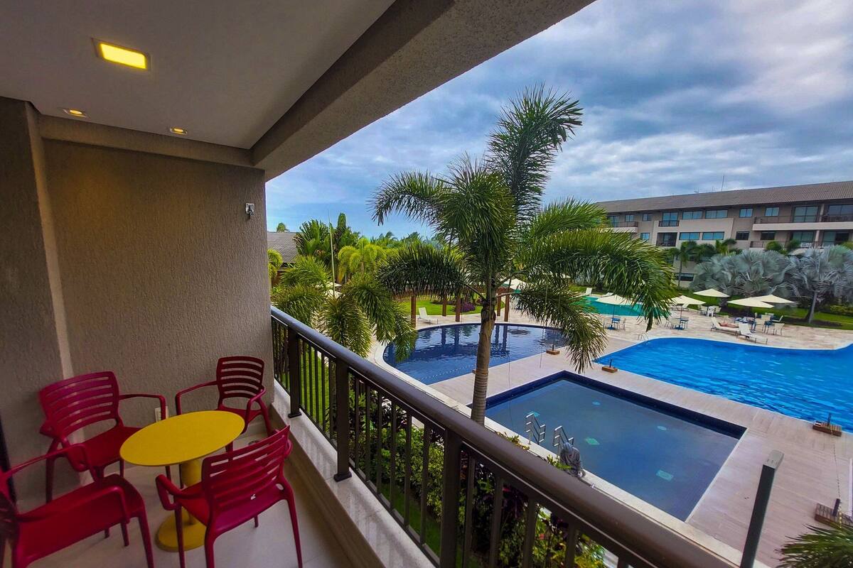 A balcony with red chairs and a yellow table, overlooking a swimming pool and a building with multiple floors.