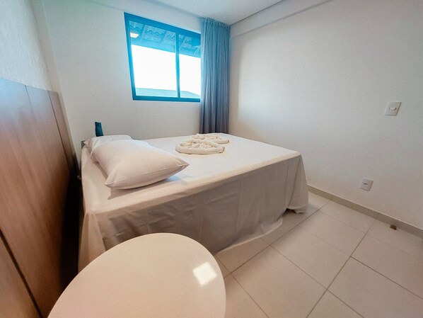 A small, clean room with a bed, a window with curtains, and a toilet. - Beach Class Residence Eco Life (Ipojuca)