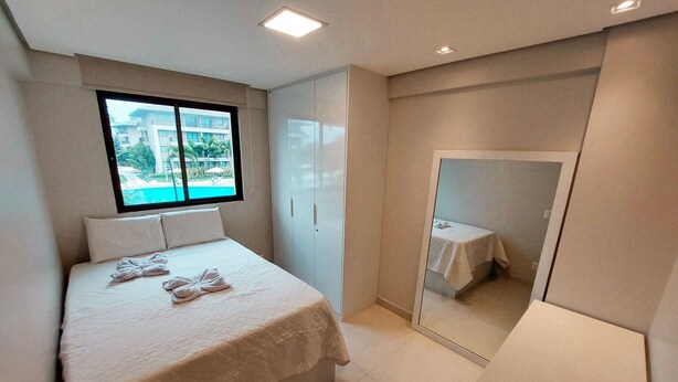A modern bedroom with a bed, a large mirror, and a window with a view of buildings and greenery. - Beach Class Residence Eco Life (Ipojuca)