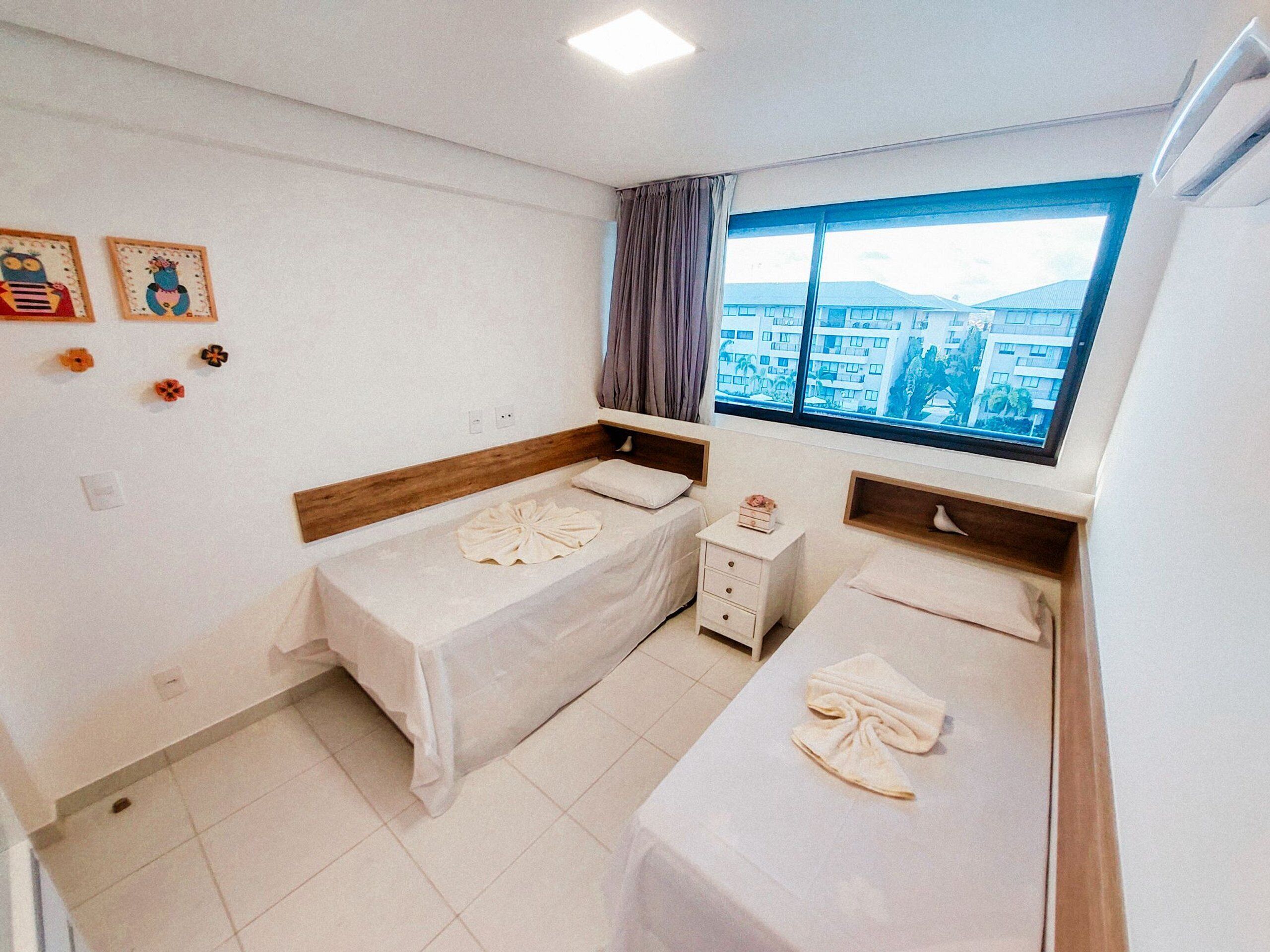 A hotel room with two single beds, a small bedside table, a window with a view of buildings, and wall decorations.