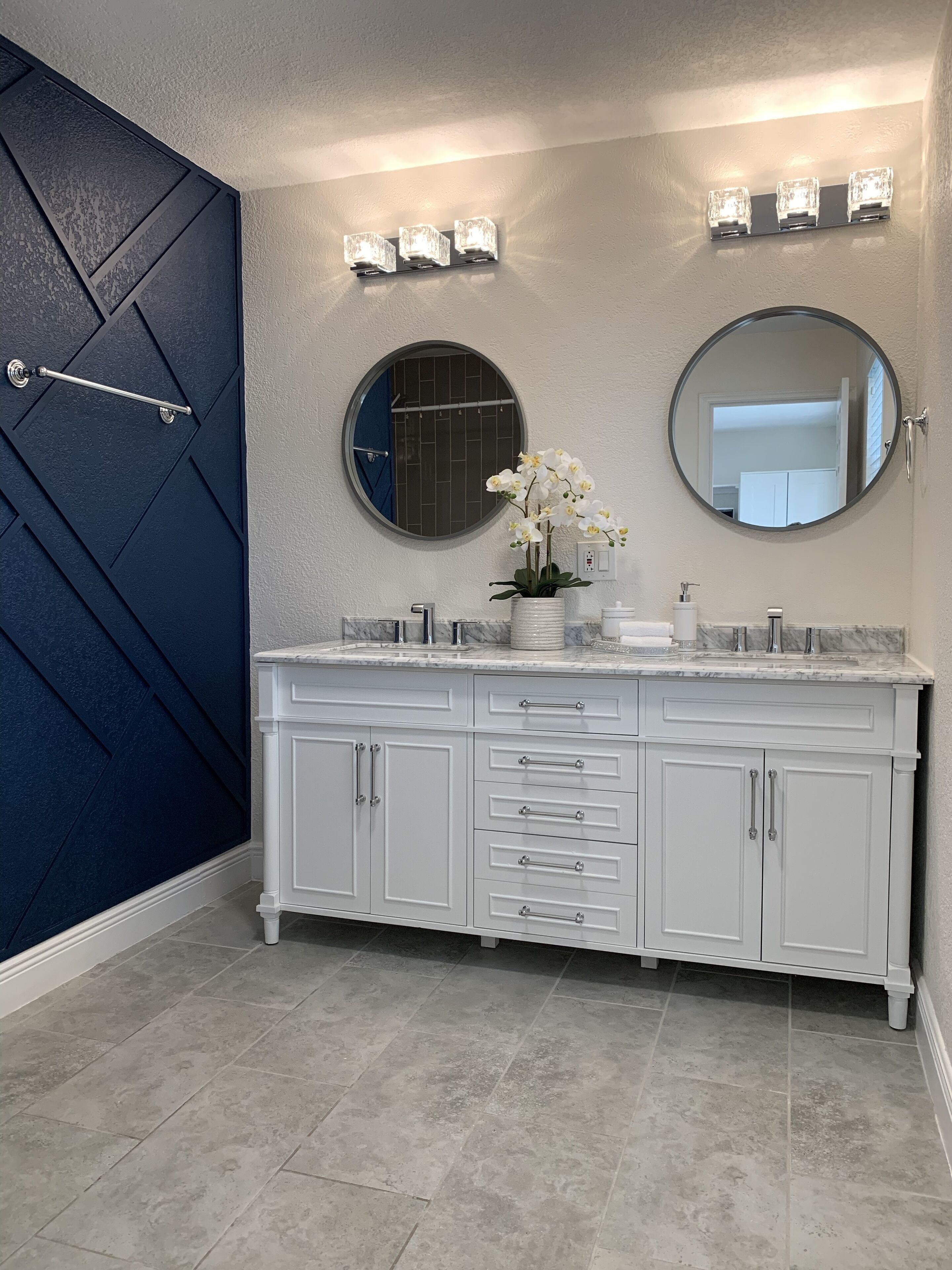 A bathroom with a dark blue door, white cabinets, a round mirror, and a potted orchid.
