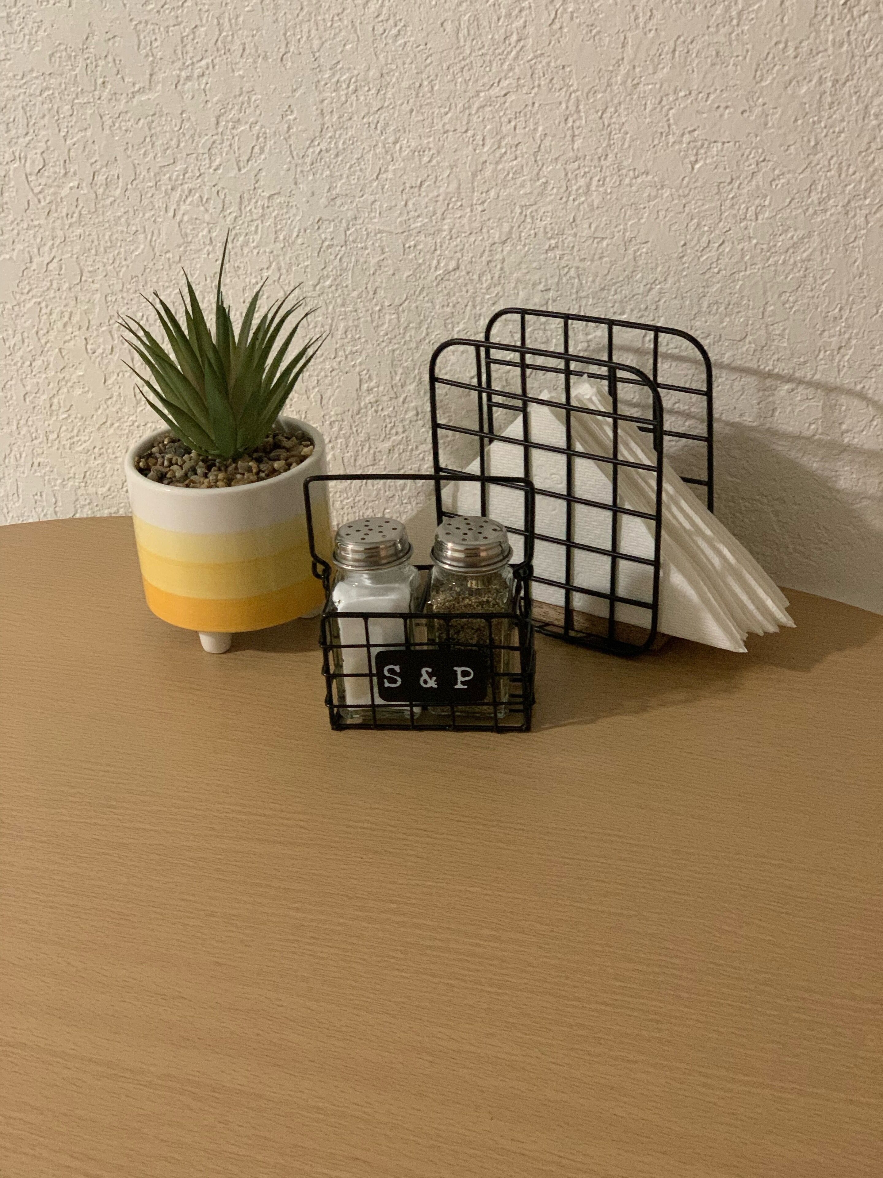 A potted plant, two salt and pepper shakers, and a black metal tray with a label on a wooden table against a textured wall.