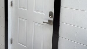 Una puerta blanca con marco negro, un candado con teclado numérico y una pequeña placa de madera con el número 1 en la pared.
