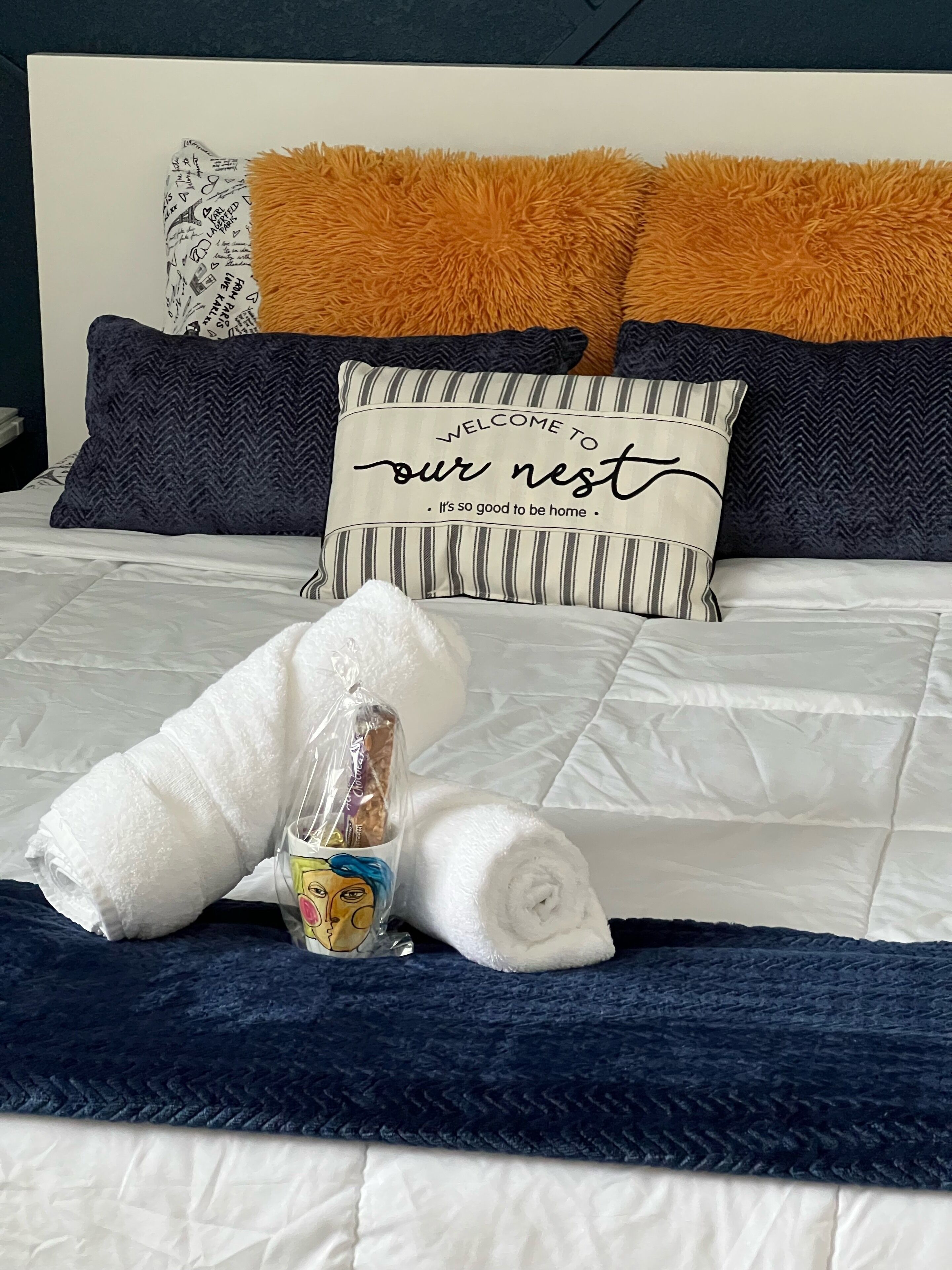 Una cama impecable con un cojín a rayas que dice 'Bienvenidos a nuestro nido. Qué bien estar en casa'.