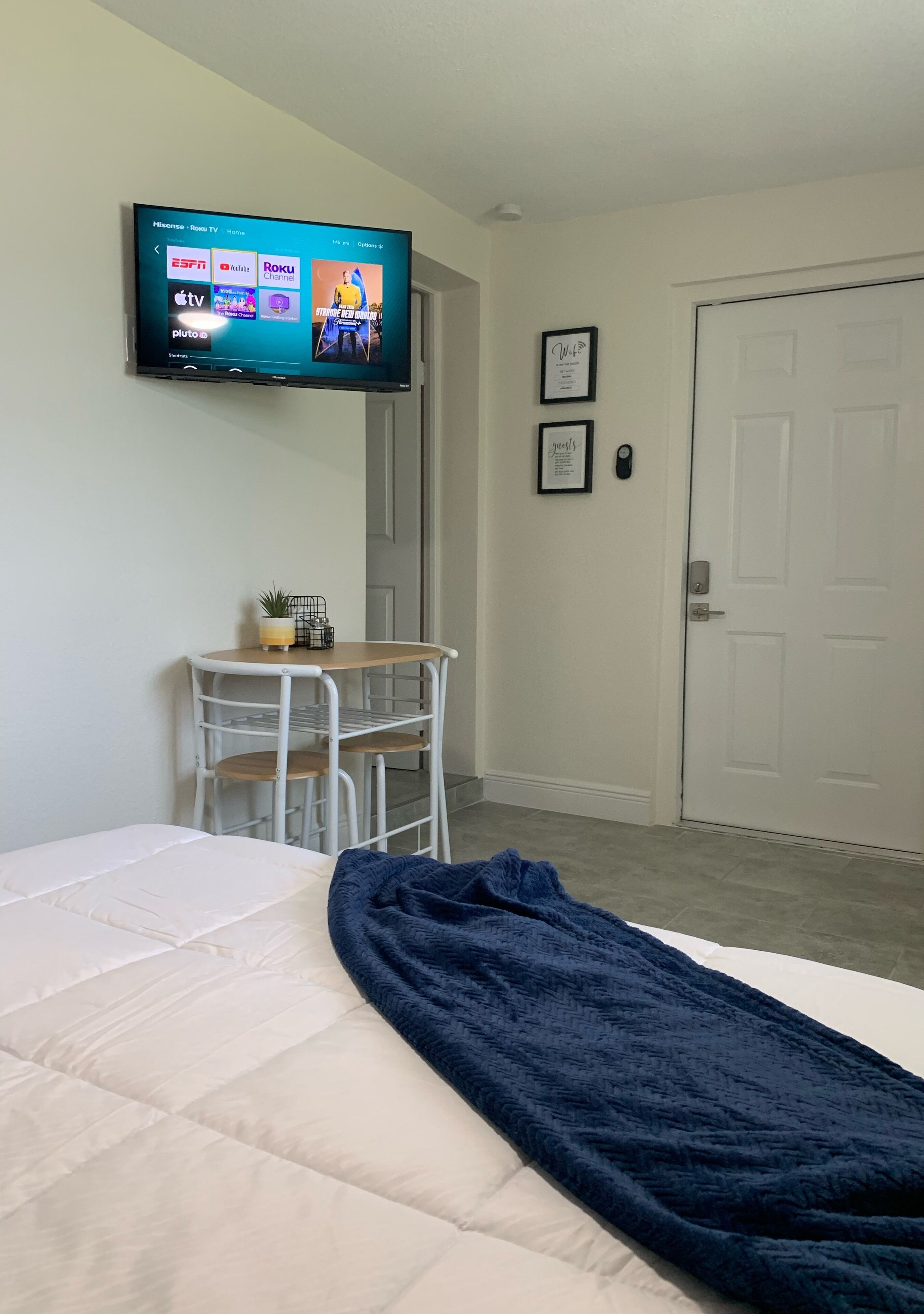 A hotel room with a bed, a TV mounted on the wall displaying various channels, a small table with a plant, and a door.