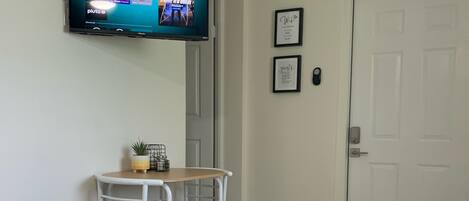 A hotel room with a bed, a TV mounted on the wall displaying various channels, a small table with a plant, and a door.
