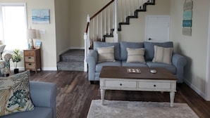 A living room with a blue sofa, a wooden coffee table, and a staircase with white railings. - Minutes to DE TURF FIELDS.  Quiet beach community.   (Frederica)