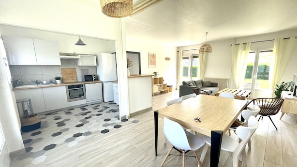 A modern kitchen with white cabinets, a dining area with a wooden table and white chairs, and a living area with a sofa and a TV.