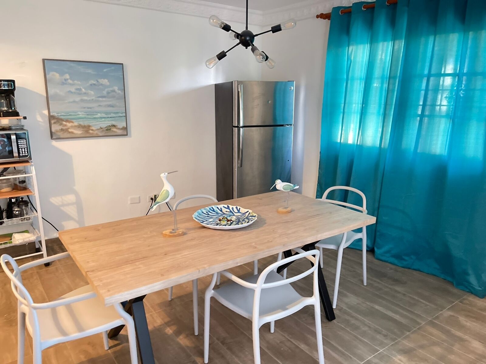Un coin repas avec une table en bois et des chaises blanches, un réfrigérateur en acier inoxydable, un luminaire fixé au mur et un rideau bleu.