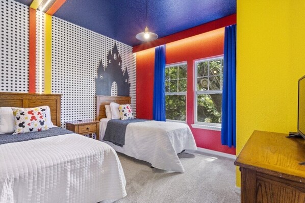 Une chambre avec deux lits, une tête de lit en bois, un téléviseur fixé au mur et une fenêtre avec des rideaux bleus.