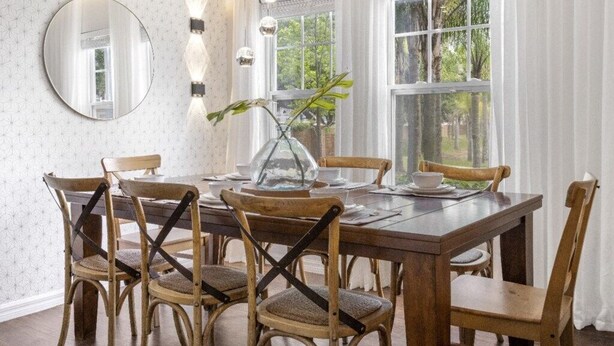 A dining room with a wooden table, six chairs, a round mirror, and a vase with a plant.