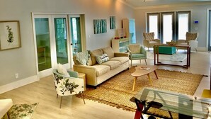 A spacious living room with a beige sofa, a green armchair, and a coffee table. There is a ceiling fan, large windows, and a view of the outdoors. - Orchid Island Family Getaway w/Private Beach and Hot Tub (Vero Beach)
