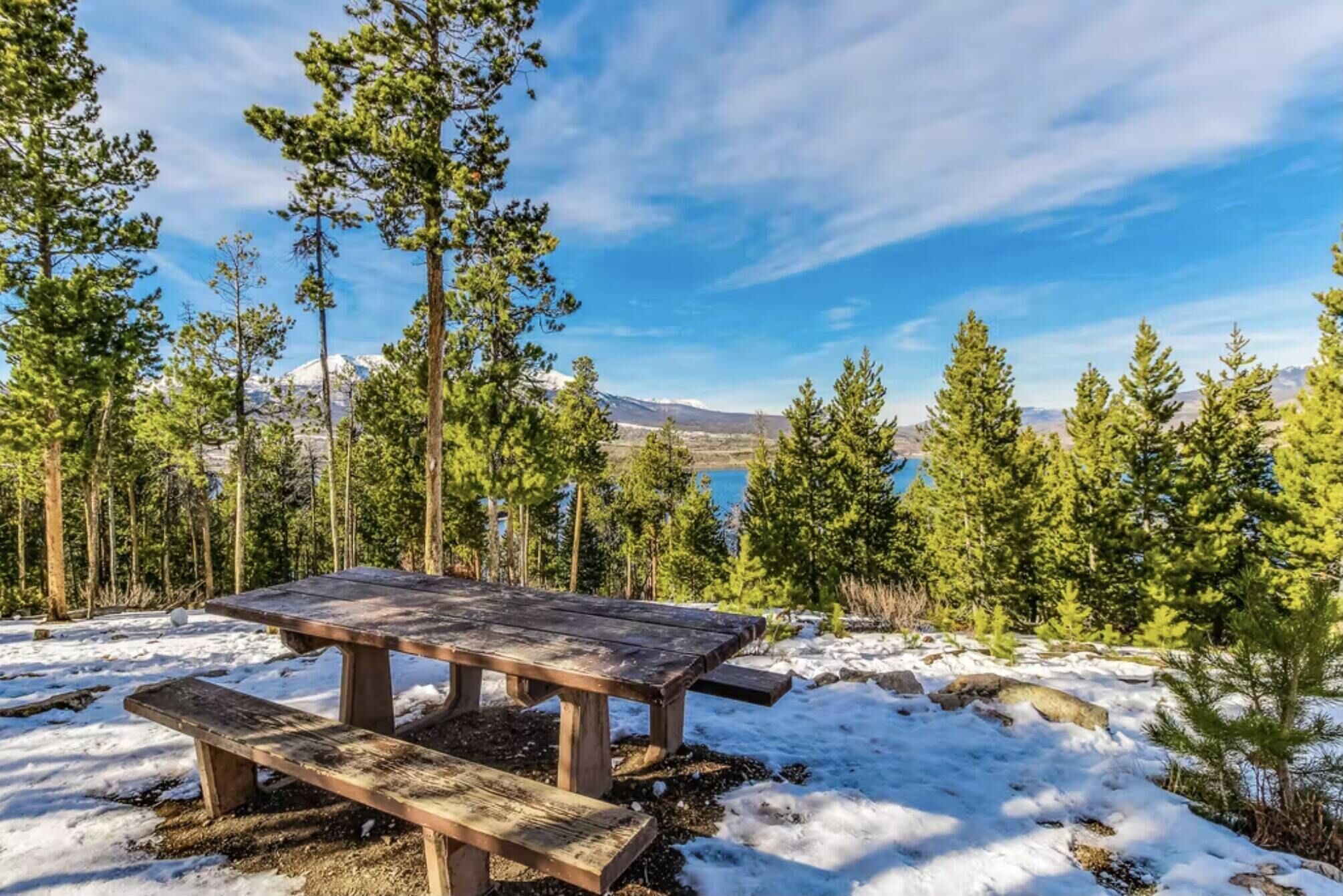 Une table de pique-nique en bois avec des bancs, située dans une forêt enneigée, avec des arbres et une montagne en arrière-plan.