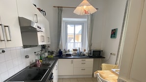 Een compacte keuken met witte kastjes, een zwart aanrecht, een raam met gordijnen en een hangende pendantlamp.