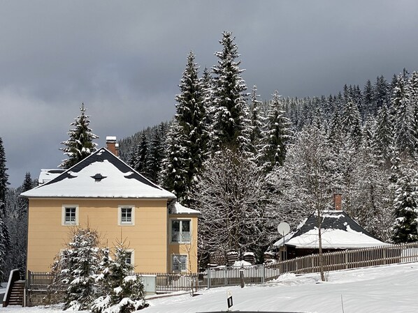 A two-story building with a snow-covered roof, surrounded by snow-laden trees and a wooden fence.
