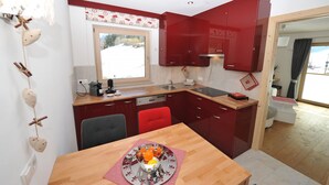 A compact kitchen with red cabinets, a wooden dining table, and a window overlooking a snowy landscape. - Apartments Leni 109A (Castelrotto)