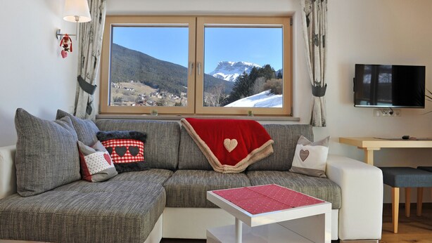 A cozy living room with a sofa, a small table, and a TV. A large window offers a view of a snowy mountain landscape. - Apartments Leni 109A (Castelrotto)