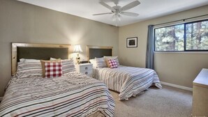 A bedroom with two beds, a ceiling fan, a nightstand with a lamp, and a window with a view of trees.