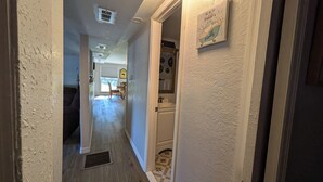 Un couloir étroit avec une porte donnant sur une salle de bain. Le couloir a un mur texturé et une grille de ventilation au plafond. On aperçoit une petite fenêtre avec des stores et une chaise au fond.