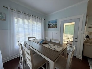 A dining area with a wooden table, white chairs, a window with white curtains, and a door leading to a balcony.
