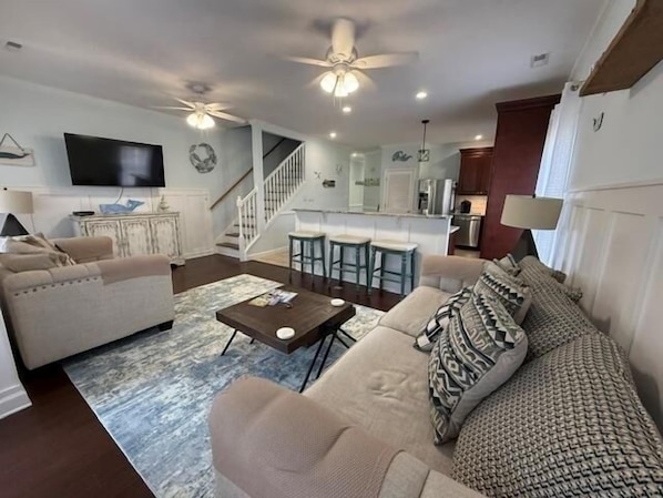 A living room with a sofa, a coffee table, and a TV. There is a staircase leading to a kitchen area.