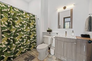 Une salle de bain avec un rideau de douche orné d’un motif de feuilles, des toilettes, un lavabo avec un miroir au-dessus et un meuble avec des tiroirs.