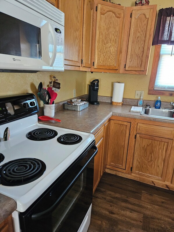 A kitchen with a white stove, a microwave, wooden cabinets, a coffee maker, and a sink. - Sweeton Creek cozy cabin just minutes from Lake Norfork (Gainesville)
