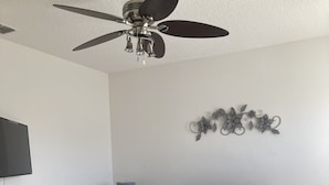 Un dormitorio con cama, ventilador de techo, televisor montado en la pared y un adorno decorativo en la pared.