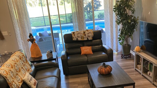 Un salón con un sofá, una mesa de centro y un mueble para televisión. Hay un ventanal con cortinas, una planta en maceta y un letrero que dice "Hocus Pocus".