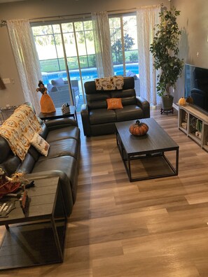 Un salon avec un canapé, une table basse et un meuble pour la télévision. Il y a une grande fenêtre avec des rideaux, une plante en pot et une pancarte sur laquelle on peut lire « Hocus Pocus ».