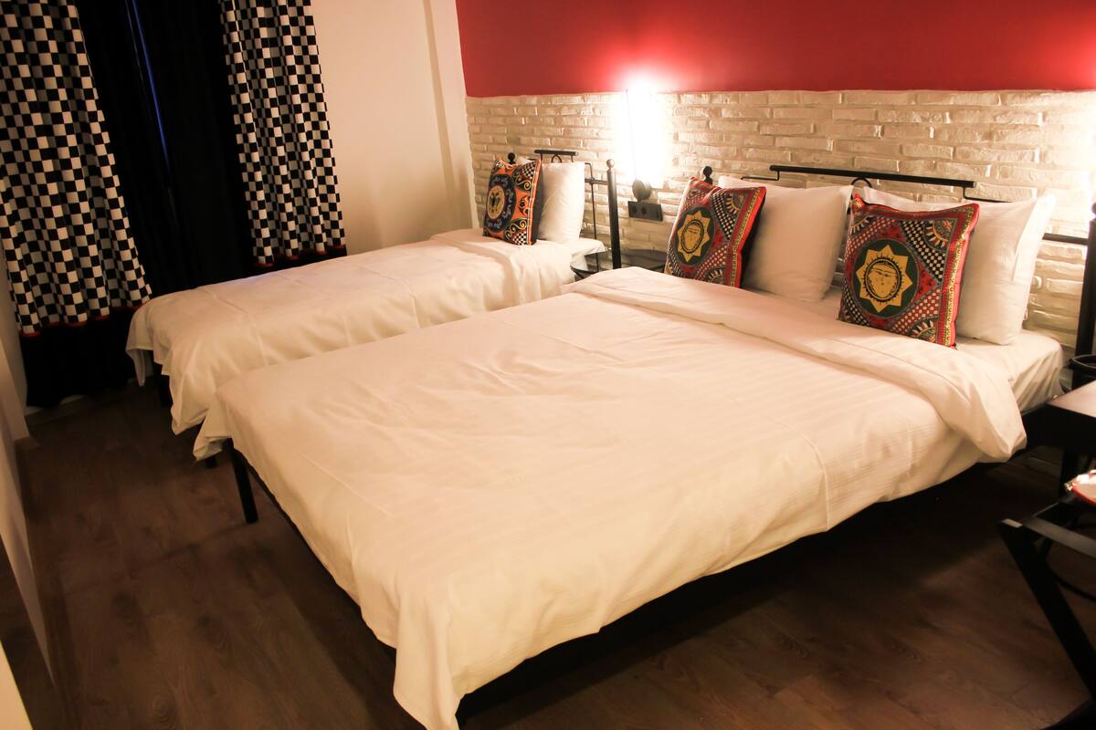 A hotel room with two beds, checkered curtains, a brick wall, and a red accent wall.