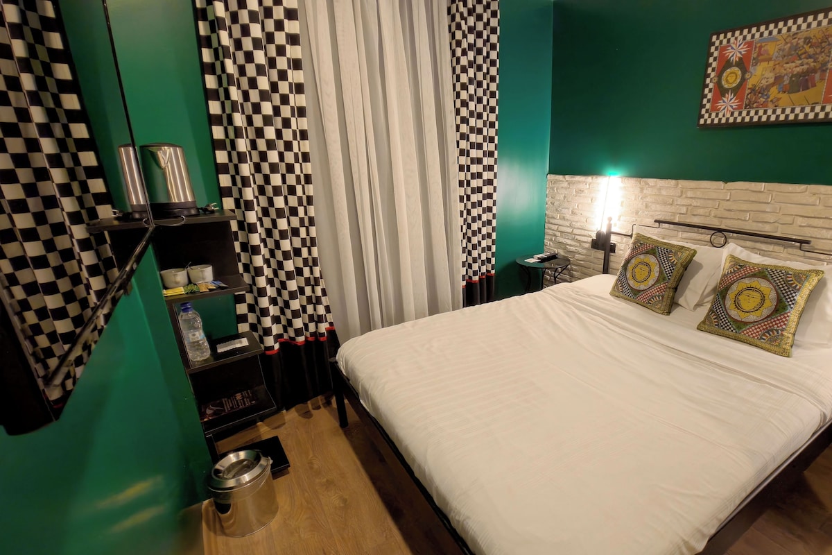 Una habitación de hotel con cortina a cuadros, una cama con almohadares estampados, una mesita y un cuadro colgado en la pared.