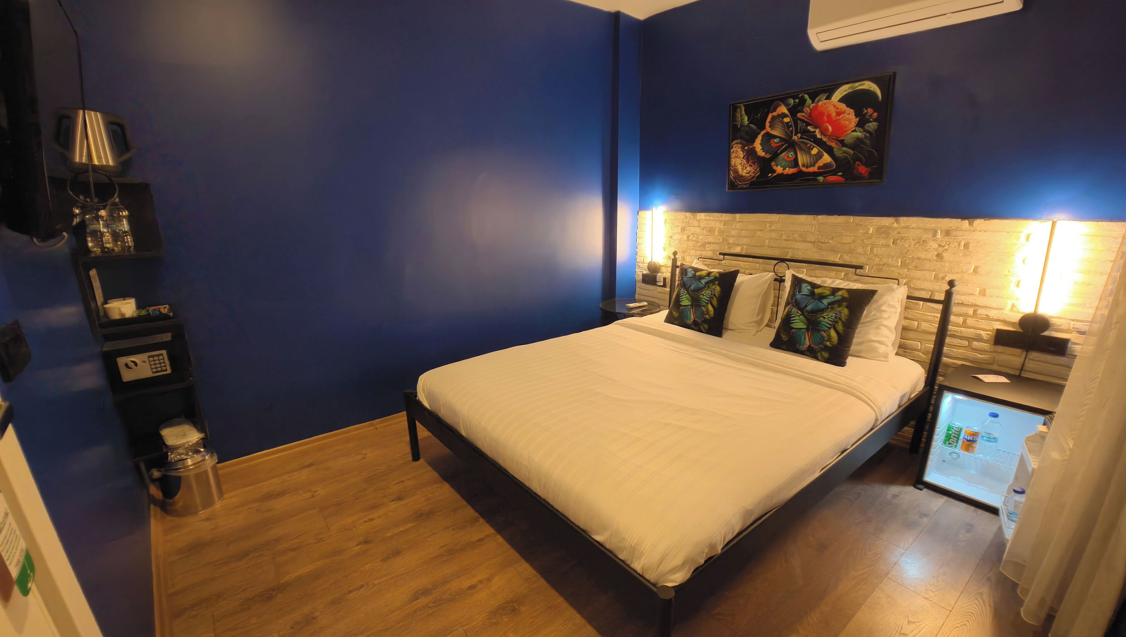 A bedroom with a bed, a nightstand, a wall-mounted TV, a small table with a lamp, and a refrigerator.