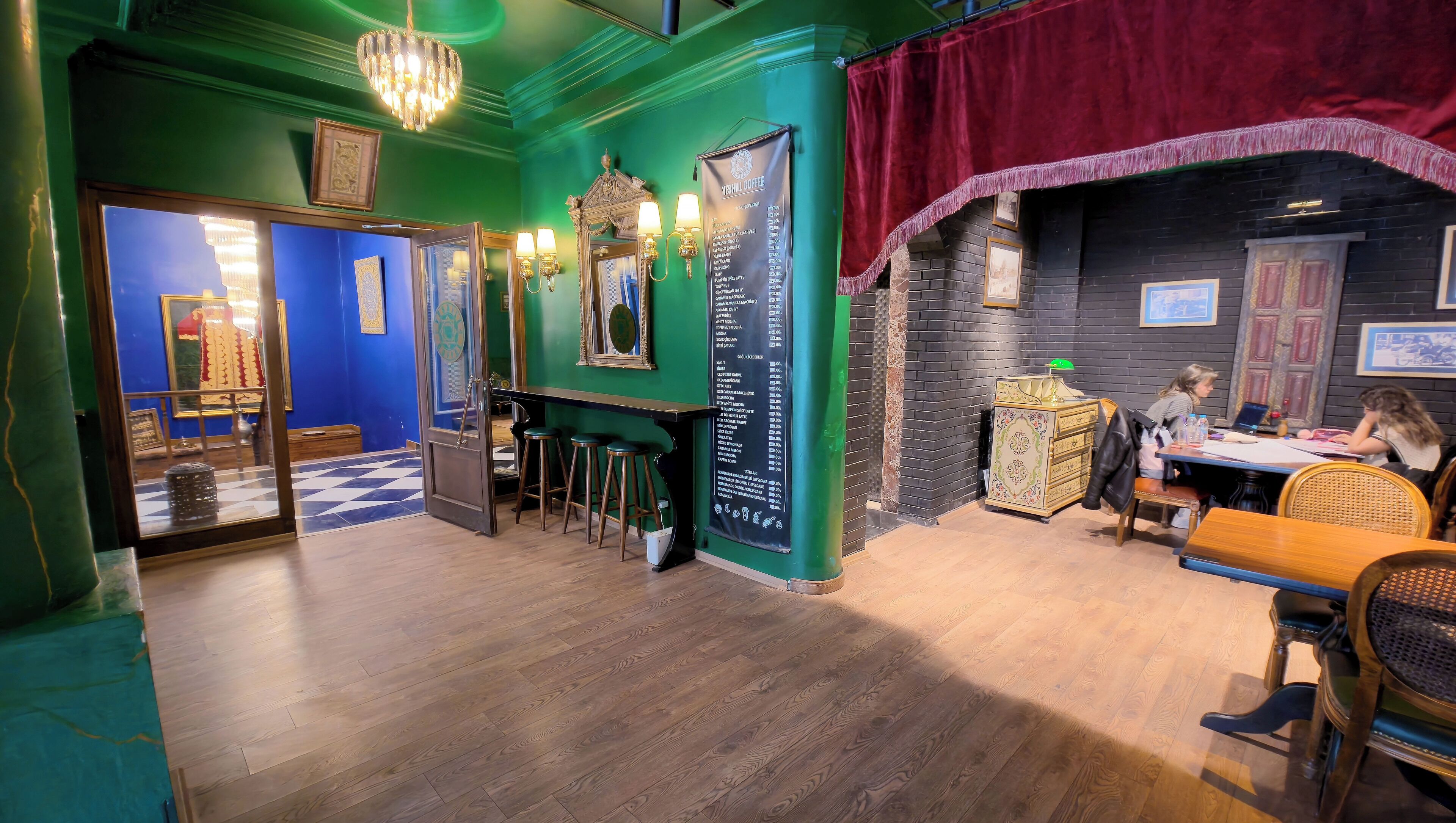 A restaurant interior with a green wall, a black brick wall, and a red curtain. There are stools and a table with people sitting.